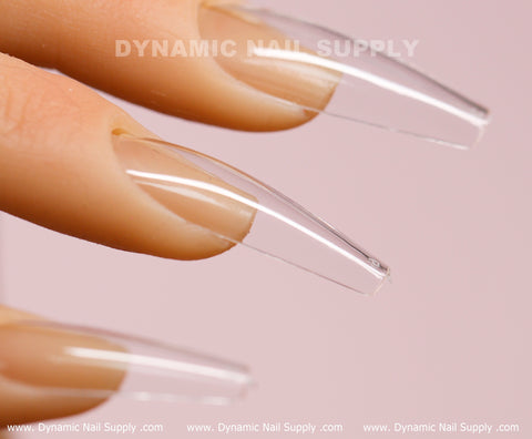 Close-up of a hand displaying elongated, glossy coffin-shaped nails using [Coffin L] Full Cover Tips - Soft Gel X tips from Dynamic Nail Supply. The clear and slightly curved acrylic extensions exude a transparent finish on natural nails. "Dynamic Nail Supply" is written across the image in white text.