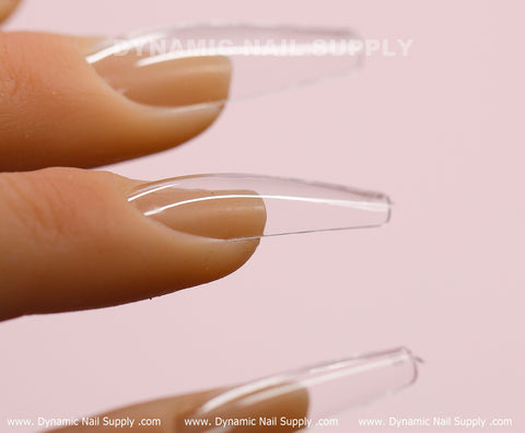 A close-up image showcases a hand with long, clear, coffin-shaped fake nails against a light pink background. The faint text at the top reads "Dynamic Nail Supply." These press-on nails are further highlighted by the repetition of www.DynamicNailSupply.com along the bottom edge. Featured in this image are Dynamic Nail Supply's [Coffin L] Full Cover Tips - Soft Gel X tips, available in a 600-piece box.