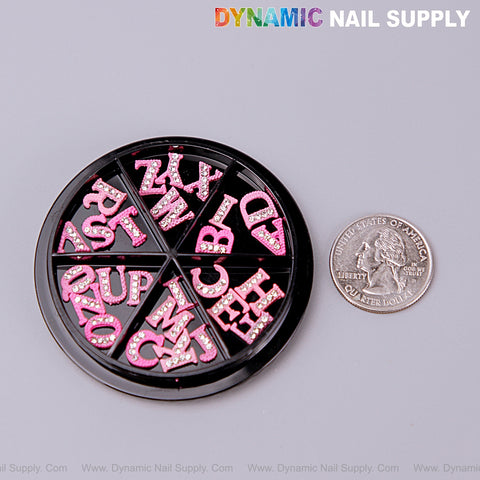 A round wheel of 26pcs pink with silver rhinestone letter nail charms A-Z shown next to a U.S. quarter for size.