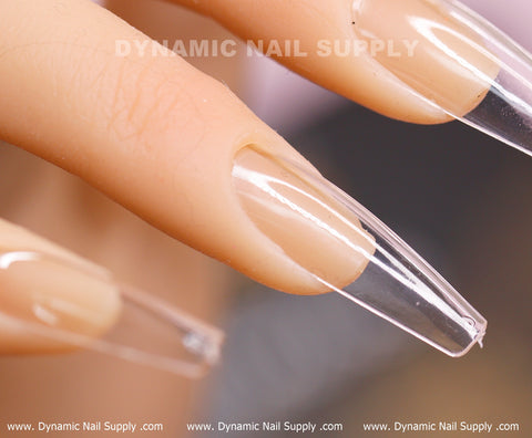 A close-up showcases a hand with elongated, clear, oval-shaped acrylic nails boasting a glossy finish. The fingers, slightly tanned and manicured, are accentuated by [Coffin L] Full Cover Tips - Soft Gel X tips from Dynamic Nail Supply. The background is blurred with the brand name prominently displayed at the top.