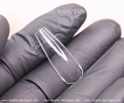 A gloved hand holds a clear, coffin-shaped nail tip from the "Coffin L" Full Cover Tips set, perfect for soft gel or full cover extensions. The transparent surface is smooth and flawless. In the background, the branding text "Dynamic Nail Supply" is artistically displayed in repetition.