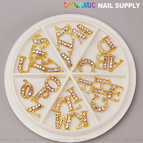A round tray holds 26 gold and silver rhinestone alphabet nail charms, A-Z, divided into lettered compartments.