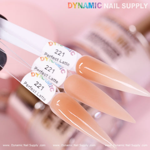 Close-up of three almond-shaped nail swatches labeled Perfect Latte - Nude Acrylic Powder (#221) from Dynamic Nail Supply, crafted with non-toxic materials. The nails flaunt a warm beige-brown hue and a glossy finish. A blurred background showcases nail bottles, with the brand name repeated along the bottom edge of the image.