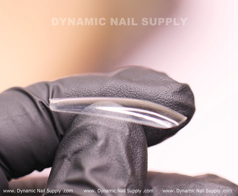 A hand in black gloves holds a transparent, elongated [Coffin L] Full Cover Tip from the Soft Gel X Tips collection, highlighting its curvature and thinness. The softly blurred background features the "Dynamic Nail Supply" logo at both the top and bottom of the image, emphasizing their premium nail products.