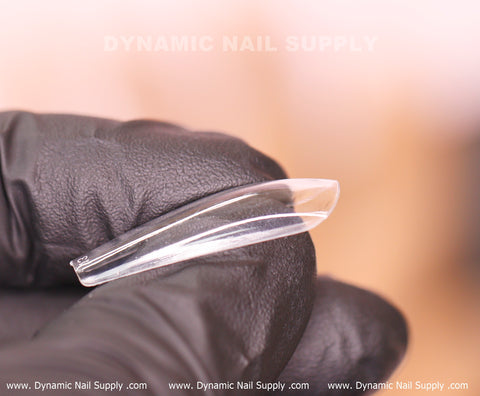 A gloved hand holds a clear, curved acrylic nail tip against a blurred background. The text "Dynamic Nail Supply" subtly appears at the top, highlighting their [Coffin L] Full Cover Tips - Soft Gel X tips - 600 pcs - Box. At the bottom, the website URL www.DynamicNailSupply.com is repeated in white letters.