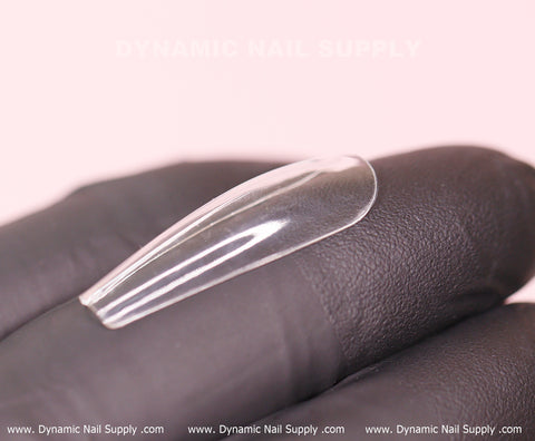 A close-up features a transparent, curved soft gel tip resting on a gloved hand, set against a light pink background. The watermark "Dynamic Nail Supply" is displayed prominently at the top, while the bottom showcases their website URL. Highlighted are premium options such as the [Coffin L] Full Cover Tips - Soft Gel X tips from Dynamic Nail Supply, available in a 600-piece box.