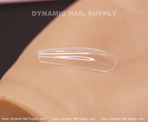 Close-up of a clear plastic false nail tip on a textured beige surface. The press-on nail, from Dynamic Nail Supply's [Coffin L] Full Cover Tips - Soft Gel X tips - 600 pcs - Box, is sharply pointed and glossy, with subtle grooves. The brand name "Dynamic Nail Supply" appears faded in the background, along with a website URL repeated along the bottom edge.