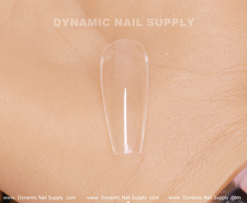 Close-up of a transparent [Coffin L] Full Cover Tip from the 600-piece Soft Gel X Tips Box resting on a skin-toned surface. The brand name, Dynamic Nail Supply, is prominently displayed at the top, and the URL www.DynamicNailSupply.com appears on a strip at the bottom.