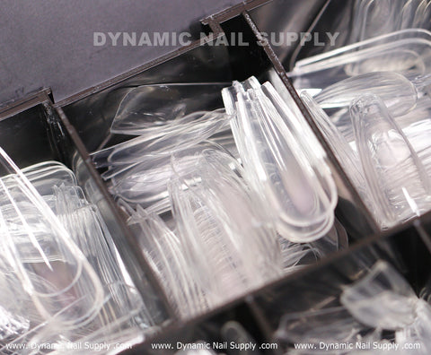 A close-up image highlights a black storage box featuring the bold white logo of "Dynamic Nail Supply." The box, labeled as [Coffin L] Full Cover Tips - Soft Gel X tips - 600 pcs, contains numerous compartments that organize an array of clear plastic full cover and soft gel nail tips. These essentials come in various sizes and shapes to accommodate different manicure styles.