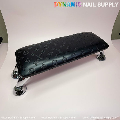 The Black LV Pattern Big Arm Rest with Leather Pillow Cushion for Manicure Nails Service by Dynamic Nail Supply features a dark embossed monogram pattern and silver chrome legs. Its plush leather pillow adds comfort and style, while the brand name is prominently displayed above in colorful and white text. The plain, light-colored background highlights the product's sleek design for an elite manicure nails service.