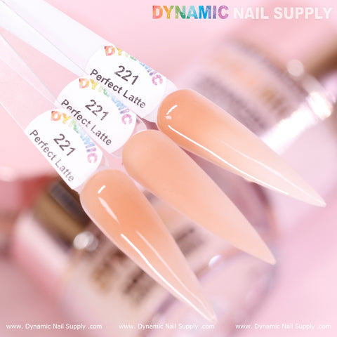 Close-up of three elongated oval nail swatches labeled as Perfect Latte - Nude Acrylic Powder (#221), showcasing a glossy, beige hue. This non-toxic acrylic powder from Dynamic Nail Supply ensures long-lasting nails. The background displays blurred nail supply items, with the colorful Dynamic Nail Supply logo at the top and a website URL at the bottom.