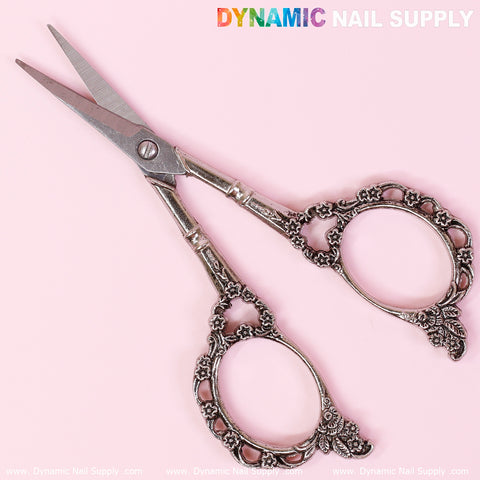 A pair of Multi-Functional Stainless Steel Nail Scissors, featuring intricate floral designs on the handles, is displayed against a light pink background. The image includes the text "Dynamic Nail Supply" in the top right corner and has a repeated watermark at the bottom.