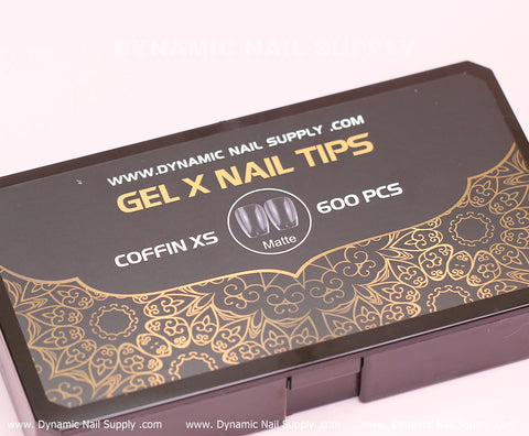 A box from Dynamic Nail Supply, featuring intricate gold patterns at the bottom edge, contains [Coffin XS] Full Cover Tips—Soft Gel X tips—in a matte finish. This black and gold package includes 600 pieces of Extra Short coffin-shaped nail tips in 11 sizes. The website www.DynamicNailSupply.com is printed at the bottom of the box.