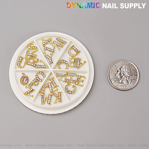 A 26pcs Gold & Silver Rhinestone Letter Nail Charms set, A-Z, shown beside a U.S. quarter for scale on gray surface.