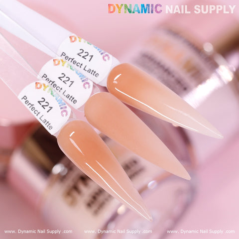 A close-up of three Perfect Latte - Nude Acrylic Powder (#221) swatches from Dynamic Nail Supply is displayed on stiletto-shaped swatch sticks. This non-toxic acrylic powder features a soft, creamy beige hue suitable for any skin tone. The blurred background showcases various nail supplies alongside the company's colorful logo.