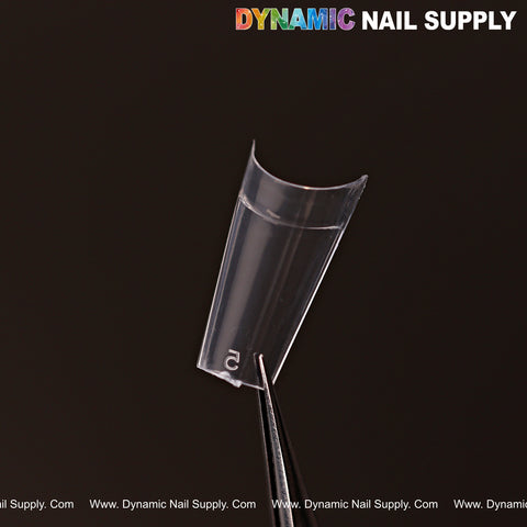 A clear short coffin nail tip from DYNAMIC NAIL SUPPLYs 600pcs 12-size set is held by tweezers on black.
