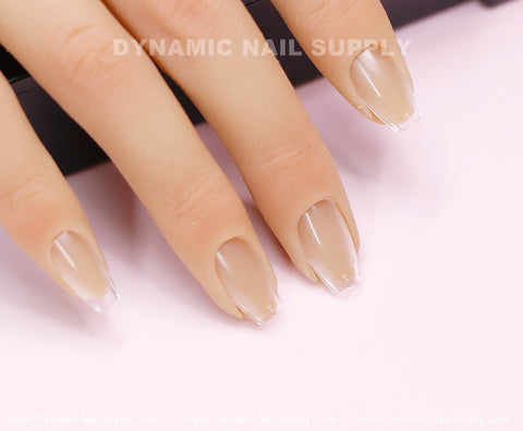 A close-up image features a hand with neatly manicured nails, highlighting a natural, translucent appearance achieved through Dynamic Nail Supply's Coffin XS Full Cover Tips from their Soft Gel X tips collection. The nails sport a light beige polish with white tips, similar to a French manicure on slightly square shapes, all set against a softly blurred background that enhances their glossy finish.