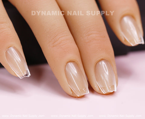 A close-up showcases fingers with coffin-shaped, pale peach Soft Gel X tips from Dynamic Nail Supply, featuring clear extensions. The skin tone is light, and the blurred background displays the brand name in white at both the top and bottom.