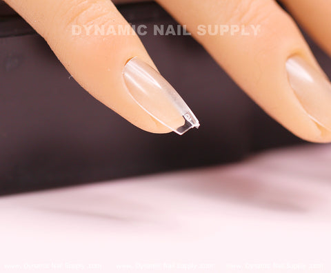 A close-up view of a practice mannequin hand highlights an extra short, coffin-shaped nail with Soft Gel X tips extension from Dynamic Nail Supply. The natural-toned nail features a partially transparent tip. In the blurred background, the words Dynamic Nail Supply can be subtly seen.