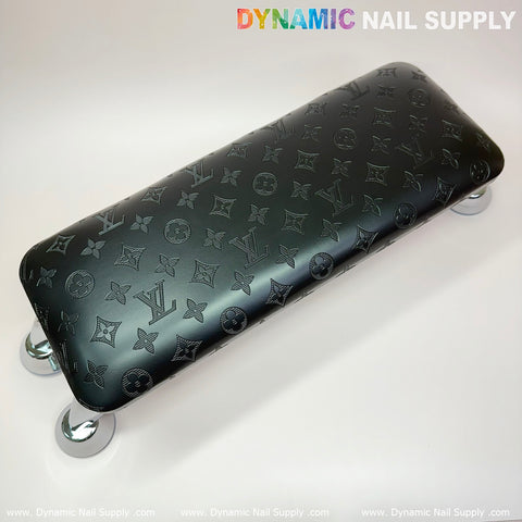 The Black LV Pattern Big Arm Rest, designed for manicure nails service, features a leather pillow cushion adorned with various logos and symbols. It is supported by four shiny metallic legs and set against a plain light background. The brand name "Dynamic Nail Supply" is prominently displayed in colorful text at the top.