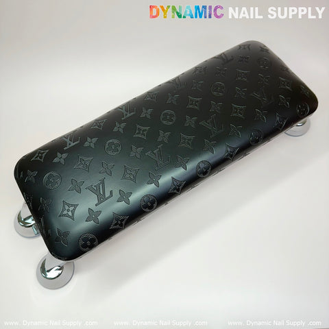 A black, rectangular leather pillow cushion with embossed LV patterns and logos is supported by four shiny, metallic legs, resembling a footrest or small bench. The plain background displays "Dynamic Nail Supply" in colorful letters along the top and bottom edges, making it perfect for enhancing the ambiance of your manicure nails service.