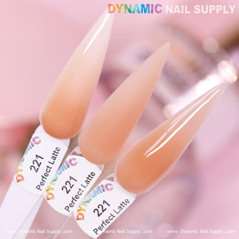 A close-up image of three elongated, pointed nail swatches showcases the Perfect Latte - Nude Acrylic Powder (#221) from Dynamic Nail Supply. Made with non-toxic materials, these swatches exhibit a glossy finish in light to medium beige tones. The labels are clearly visible against a softly blurred, pink-toned background that highlights the elegance of the nail colors.