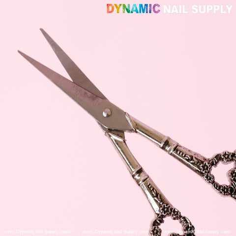 Close-up of the Multi-Functional Stainless Steel Nail Scissors by Dynamic Nail Supply with intricate handles on a pink background. The slightly open stainless steel blades gleam sharply, with detailed engravings enhancing their elegance. The top right features "Dynamic Nail Supply" in bold, colorful text.