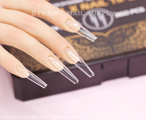 A mannequin hand showcases extra-long, clear coffin-shaped nail extensions, elegantly presented on a black box adorned with gold detailing and partially inscribed with Dynamic Nail Supply. Set against a soft pink background, the beautifully manicured nails shine with modern sophistication, thanks to the durable Coffin XXL Full Cover Tips from Dynamic Nail Supply's Soft Gel X tips collection.