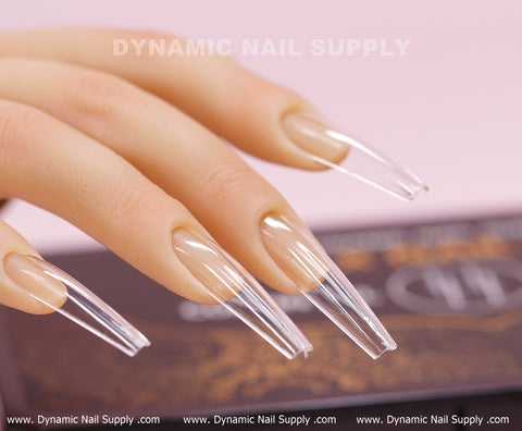 A close-up of a hand featuring long, clear [Coffin XXL] Full Cover Tips from Dynamic Nail Supply, shaped in an extra-long coffin style. These sharply pointed Soft Gel X tips offer a polished and glossy transparent appearance. In the blurred background, there's a sign for Dynamic Nail Supply set against a soft pink backdrop.