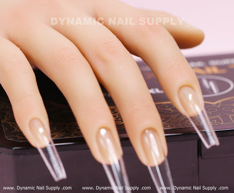 Close-up of a hand showcasing glossy, extra-long coffin-shaped nail extensions with French tips, gracefully placed on a black box embellished with gold patterns. The [Coffin XXL] Full Cover Tips - Soft Gel X tips exhibit a contemporary, expertly manicured look. Subtle branding in the background features Dynamic Nail Supply.