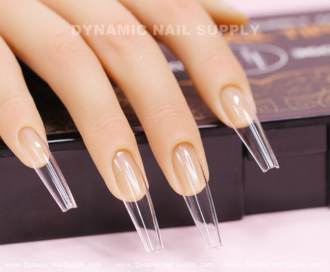 Close-up of a hand showcasing Coffin XXL full cover tips in extra long, coffin shape atop a mannequin. The translucent soft gel X tips accentuate their sharp, elongated form. In the background, a blurred box labeled with the brand name Dynamic Nail Supply adds context to the scene.