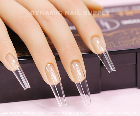 A model hand displaying extra long, clear full cover soft gel nails in a coffin shape, named [Coffin XXL], shows off their glossy and sharply tapered design. Behind the nails is a black and brown box with subtle text, while the surface beneath features light pink branding from Dynamic Nail Supply.