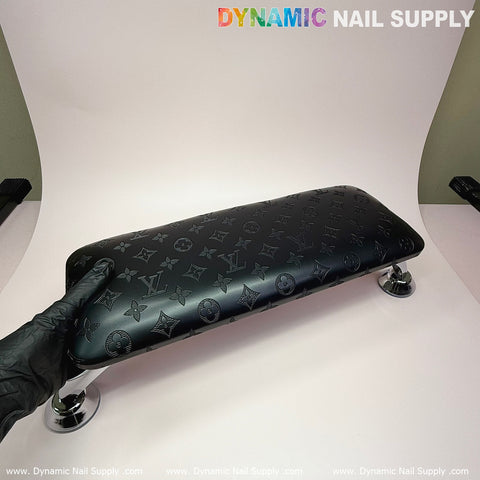 A (Black LV Pattern) Big Arm Rest with a leather pillow cushion for manicure services, featuring a glossy finish and branded motifs, rests on a white surface next to a sleek UV nail light. Chrome legs offer elegant support as someone wearing black gloves adjusts it. The background showcases the distinctive Dynamic Nail Supply label.