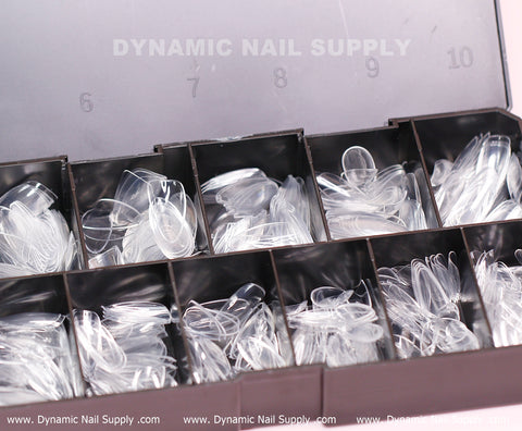 A plastic organizer box with multiple compartments contains a selection of Almond M Full Cover Tips, crafted by Dynamic Nail Supply. These soft gel X tips come in a set of 600 pieces, covering 10 sizes in a medium long shape. The lid bears the Dynamic Nail Supply logo, and the base prominently displays the repeated website address. Each tip is meticulously sorted and ready for use.