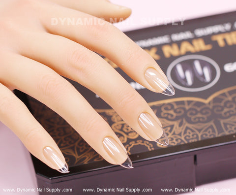 A hand adorned with long, clear, almond-shaped press-on nails from the [Almond M] Full Cover Tips by Dynamic Nail Supply is displayed on a black and gold box labeled Dynamic Nail Supply. These Medium Long Soft Gel X tips boast a glossy finish, showcasing intricate patterns against the soft pink background.