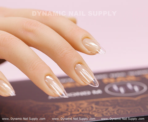 Close-up of a hand showcasing almond-shaped, clear acrylic nails accentuated by the [Almond M] Full Cover Tips from Dynamic Nail Supply. The glossy, well-manicured nails are highlighted against a backdrop of intricate golden patterns on a dark surface, with the Dynamic Nail Supply brand name subtly appearing at the top.