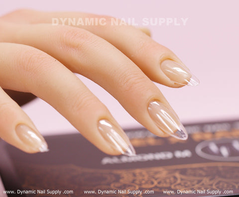 A close-up shot showcases a mannequin hand adorned with [Almond M] Full Cover Tips on a pink background. The transparent Soft Gel X tips from Dynamic Nail Supply glisten with detailed artistry. Both the brand name, Dynamic Nail Supply, and a patterned box are visible in the backdrop and foreground.