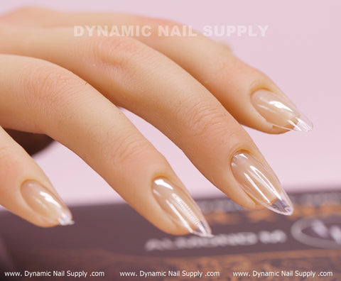 The image showcases a detailed view of a hand adorned with Dynamic Nail Supply's [Almond M] Full Cover Tips, featuring 600 medium-long, glossy nude Soft Gel X nail tips in a medium almond shape. The background displays blurred pink hues, and "Dynamic Nail Supply" is prominently featured at the top.