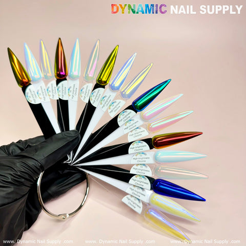 A gloved hand holds a ring of elongated, pointed nail samples in various metallic and iridescent colors, including vibrant hues like gold, silver, blue, and red. The stunning array is enhanced with Orange/Gold/Mermaid Iridescent Chrome #06 - High Brightness Fine Pigment Powder for extra shine, showcasing remarkable nail art potential. The brand name Dynamic Nail Supply is prominently displayed at the top against a soft pink background.