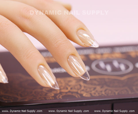 A hand displays medium long, almond-shaped, clear Full Cover Tips from the [Almond M] Soft Gel X collection by Dynamic Nail Supply on a decorative surface. The glossy finish highlights the natural nail beneath. In the background, Dynamic Nail Supply's logo is visible alongside repeated website URLs. These stylish tips perfectly imitate elegant design aesthetics.