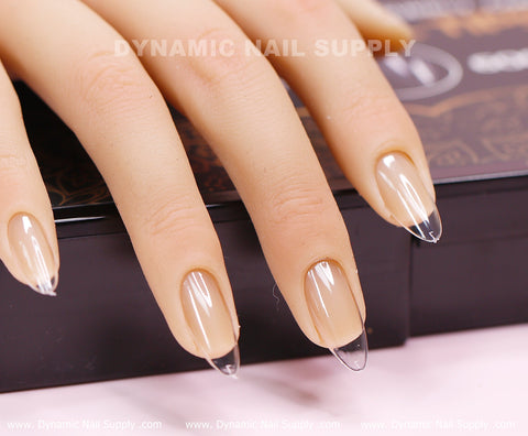 Close-up of a hand with well-manicured nails featuring clear, pointed [Almond M] Full Cover Tips in a Medium Almond Shape. The natural beige base transitions elegantly to a transparent tip. The hand rests on a dark surface, with the Dynamic Nail Supply brand subtly in the background.