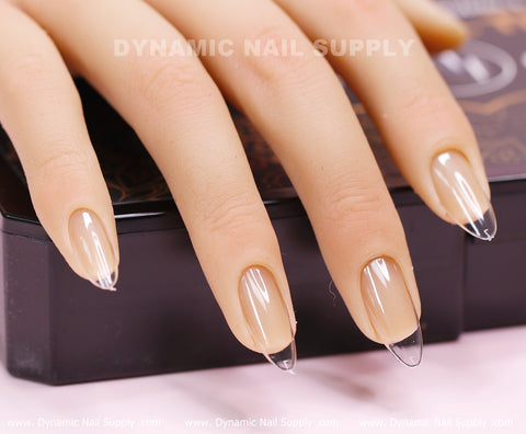 A close-up shot captures a hand adorned with soft gel "Almond M" medium-long tips from Dynamic Nail Supply, showcasing their almond shape with clear, pointed design and beige nail beds. The hand is placed on a black surface where the brand name, Dynamic Nail Supply, is visible in the background.