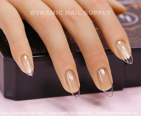 A close-up of a mannequin hand displays long, almond-shaped acrylic nails created using the Almond M Full Cover Tips from Dynamic Nail Supply. The fingertips rest casually on a black surface, with blurred text in the background subtly highlighting the brand.