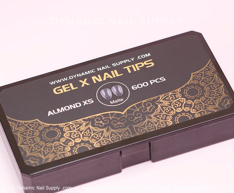 A black box with an intricate gold design holds 600 Full Cover Tips from Dynamic Nail Supply, ideal for creating Extra Short Almond Shape nails with a matte finish. The box is labeled as DYNAMIC NAIL SUPPLY, GEL X NAIL TIPS, ALMOND XS, and features the website DYNAMICNAILSUPPLY.COM at the top.