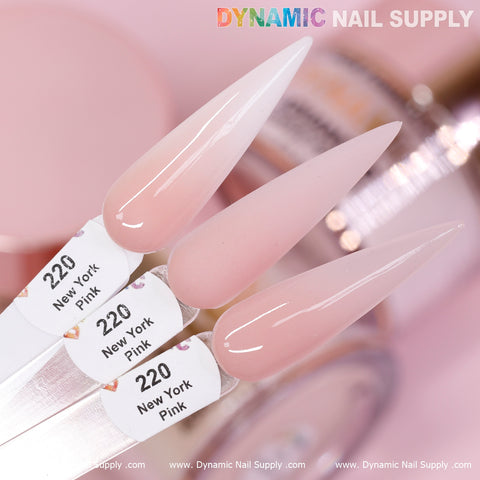 Close-up of three nail polish swatches labeled New York Pink - Nude Acrylic Powder (#220) from Dynamic Nail Supply. These long-lasting polishes showcase a spectrum from soft white to light pink. The background subtly displays blurred containers of similar shades, enhancing a cohesive pastel aesthetic.