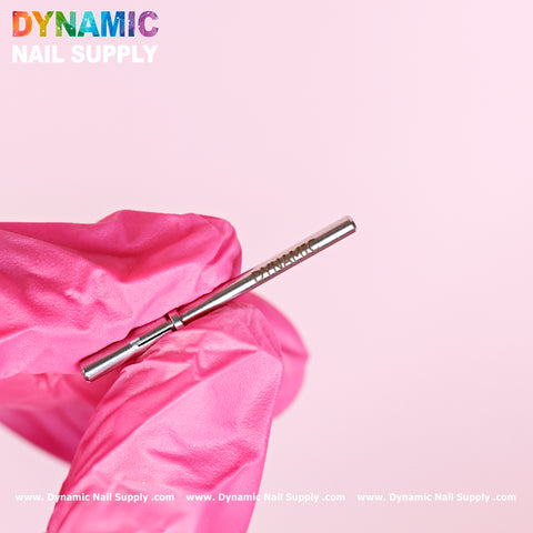 A hand wearing a vibrant pink glove carefully holds a sleek, metallic nail drill bit with the Small 3.5mm Easy-Off Mandrel for Smaller Sanding Band against a gentle pink backdrop. The brand name Dynamic Nail Supply appears in multicolored text at the top left corner, and the website address is displayed in white at the bottom edge of the image.