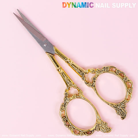 The Multi-Functional Stainless Steel Nail Scissors from Dynamic Nail Supply, featuring elegant floral details on the handles, rest against a soft pink background. The ornate design provides a vintage touch reminiscent of fine embroidery work, while the sleek blades are slightly open. The top displays "Dynamic Nail Supply" in colorful, bold letters.