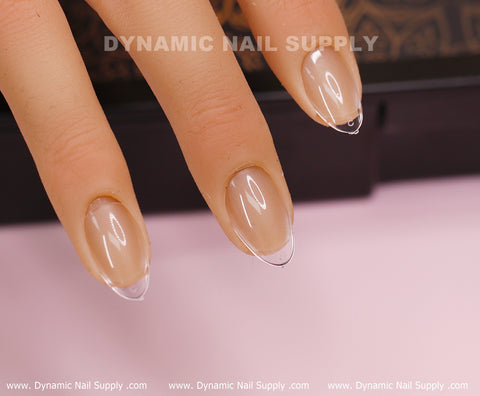 A close-up of a mannequin hand showcases clear, extra short almond-shaped artificial nails over a natural beige base using Almond XS Full Cover Tips by Dynamic Nail Supply. The background is a soft pink, and the text "DYNAMIC NAIL SUPPLY" is displayed both on the nails and along the lower border.