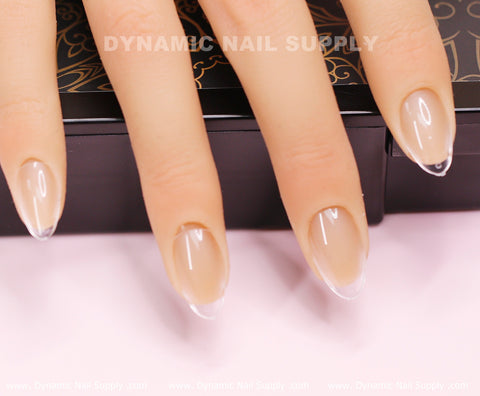 The image features a close-up of a hand with elegantly manicured nails, polished in light beige with clear tips. The nails display an extra short almond shape and rest against a decorative black surface. In the background, a subtle design includes text highlighting Dynamic Nail Supply's [Almond XS] Full Cover Tips - Soft Gel X tips, which are available in 11 sizes for added style.