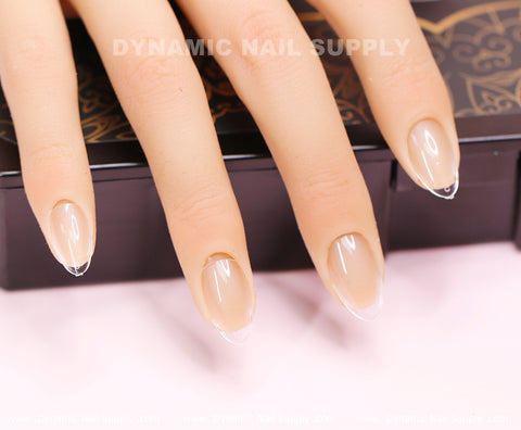 A close-up highlights a hand flaunting nails adorned with the [Almond XS] Full Cover Tips by Dynamic Nail Supply, showcasing an extra short almond-shaped design. These Soft Gel X tips feature a natural nude base with a clear, glossy tip reminiscent of a modern French manicure. Elegant black and gold patterns in the background enhance the scene's sophistication.